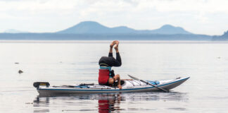 man performs a headstand while kayaking in calm waters with mountains in background
