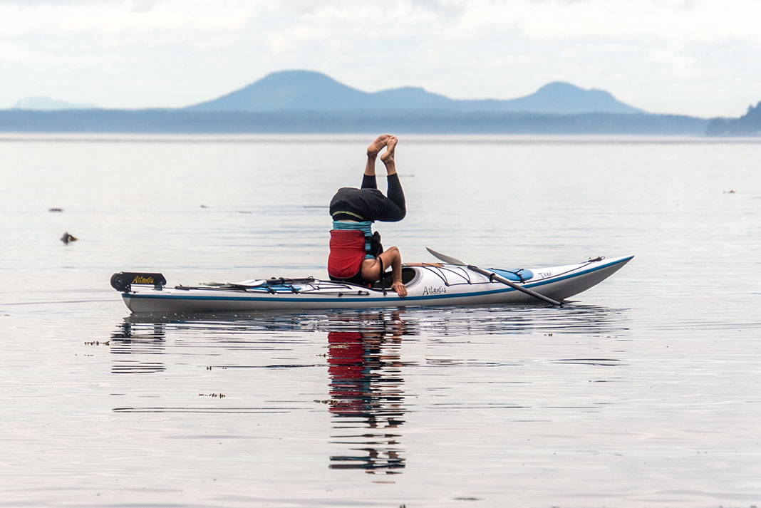 man performs a headstand on a kayak in calm waters with mountains in background, and his head is sticking inside the cockpit