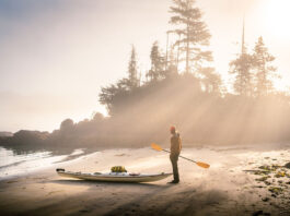 man stands on beach holding paddle before sea kayaking, thinking about Olympic events