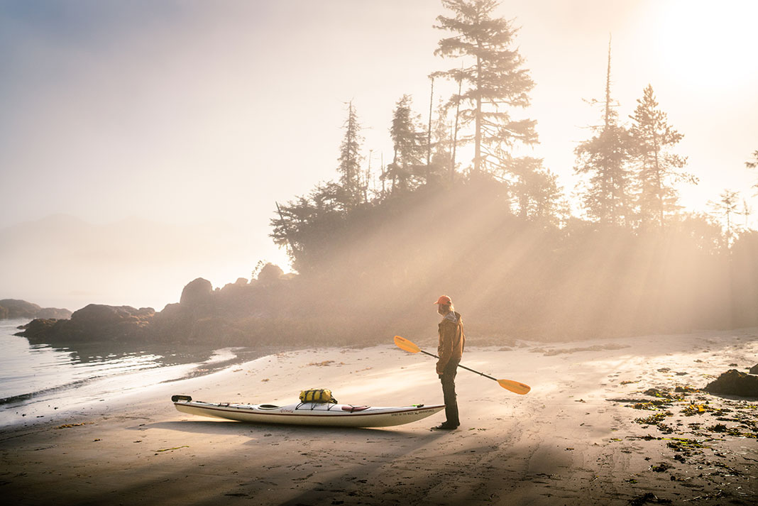 man stands on beach holding paddle beside a sea kayak