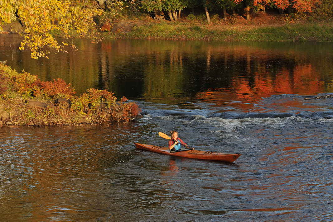 woman paddles a wooden kayak in front of fall foliage
