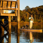 Hybrid SUP Kayak Review: Hobie Mirage Lynx man stands and casts near a pier in the Hobie Mirage Lynx