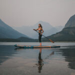 Best Body Glove Paddleboards For 2026 Woman on inflatable paddleboard on lake with mountain in background