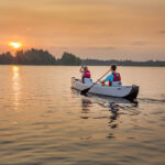Best Inflatable Canoes For 2026 Two people paddling inflatable canoe towards the setting sun on a lake