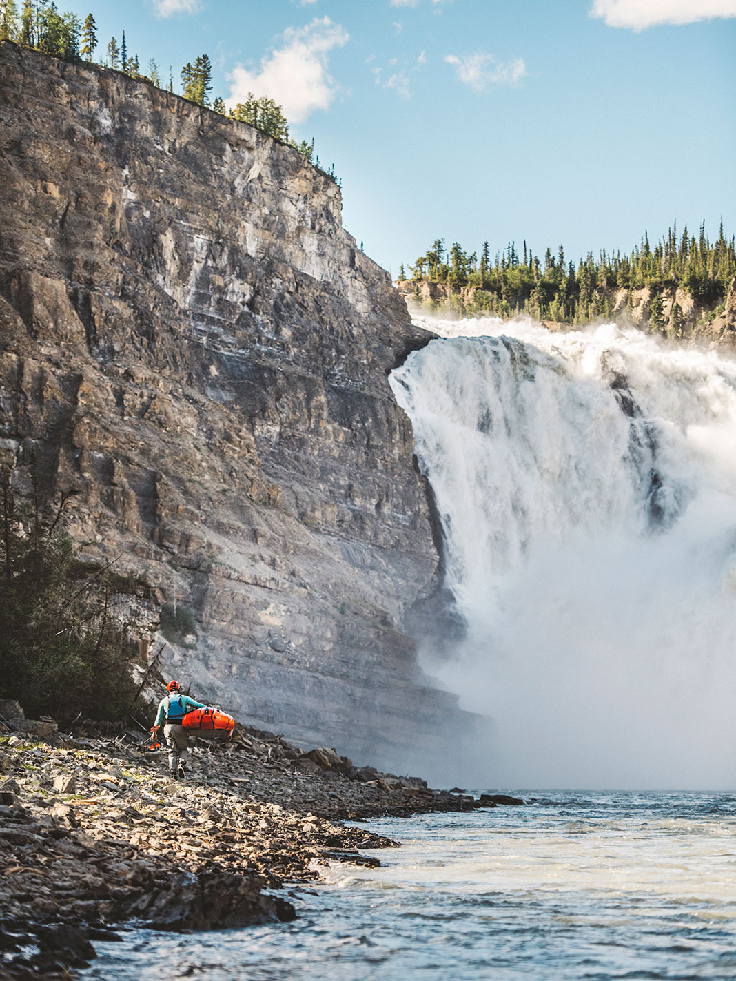 person carries a whitewater kayak back toward a very large waterfall on the Nahanni River
