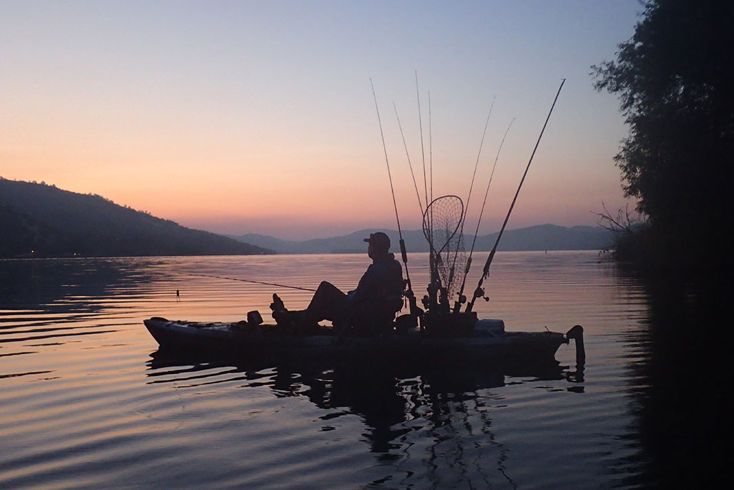 kayak angler pedals across calm water at dusk with California mountains in the background