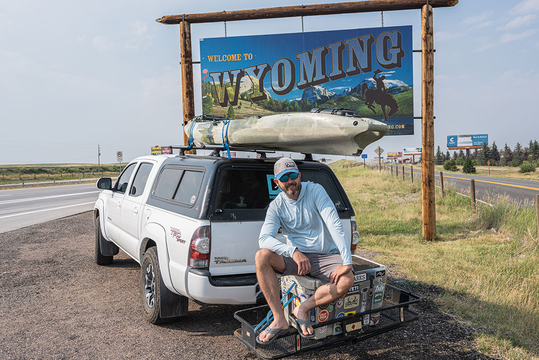 man with white truck carrying fishing kayak poses in front of Wyoming sign