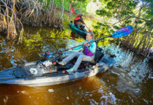 a woman and man paddle their pedal kayaks through tight quarters on a muddy river