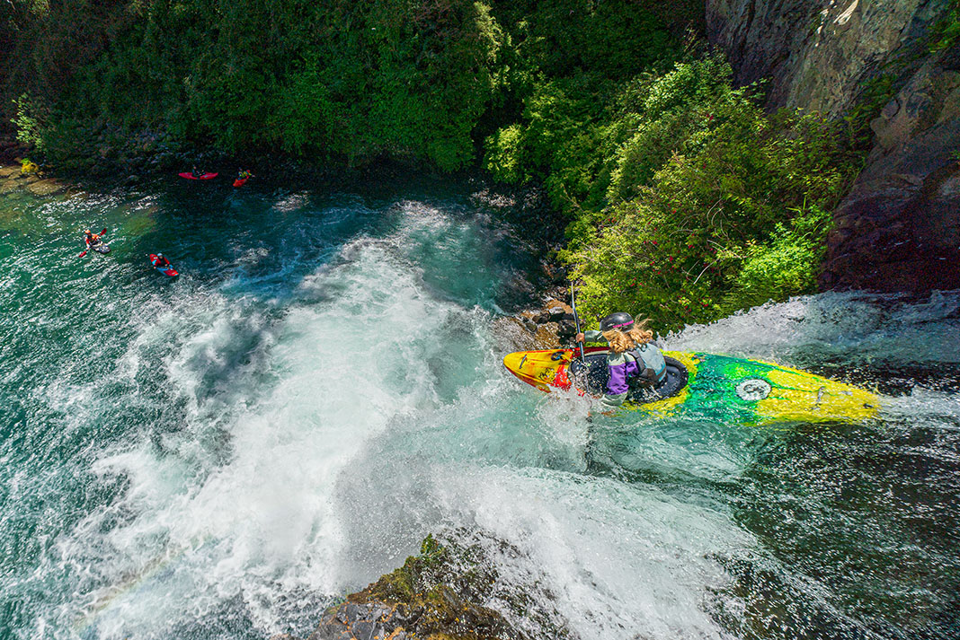 a female whitewater kayaker runs a waterfall while her paddling crew watches from below