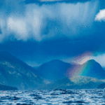 The Love And Madness Of Long-Distance Expeditions a long distance expedition kayaker paddles the cloudy Kyuquot Crossing in front of a rainbow