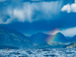 a long distance expedition kayaker paddles the cloudy Kyuquot Crossing in front of a rainbow