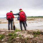 Back In The Pack: Ostrom Outdoors Returns a man and woman stand beside a northern river wearing Ostrom Outdoors canoe packs