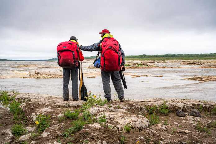 a man and woman stand beside a northern river wearing Ostrom Outdoors canoe packs