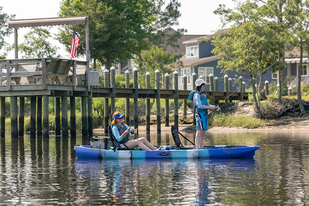 two women sit and stand while fishing from a blue tandem Brooklyn Kayak Company TK122 fishing kayak in front of a pier with American flag