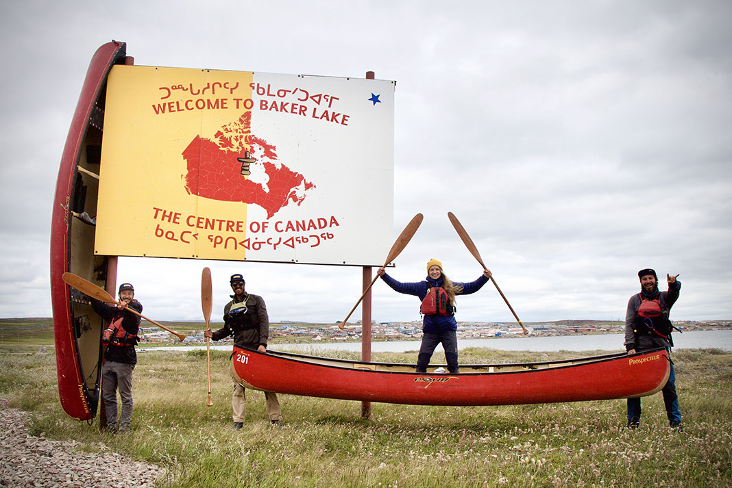 a group of expedition canoeists pose with two red canoes in front of the Baker Lake sign