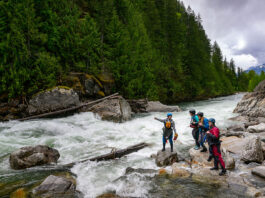a group of female whitewater paddlers scout a river while wearing womens drysuits
