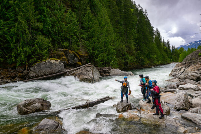 a group of female whitewater paddlers scout a river while wearing womens drysuits
