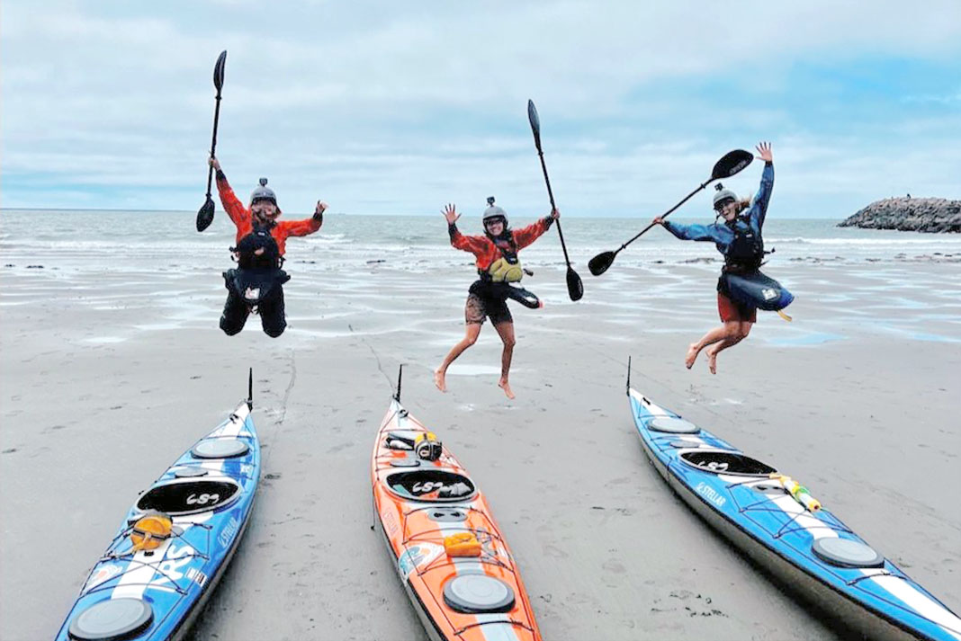 three female sea kayakers jump in front of their beached sea kayaks
