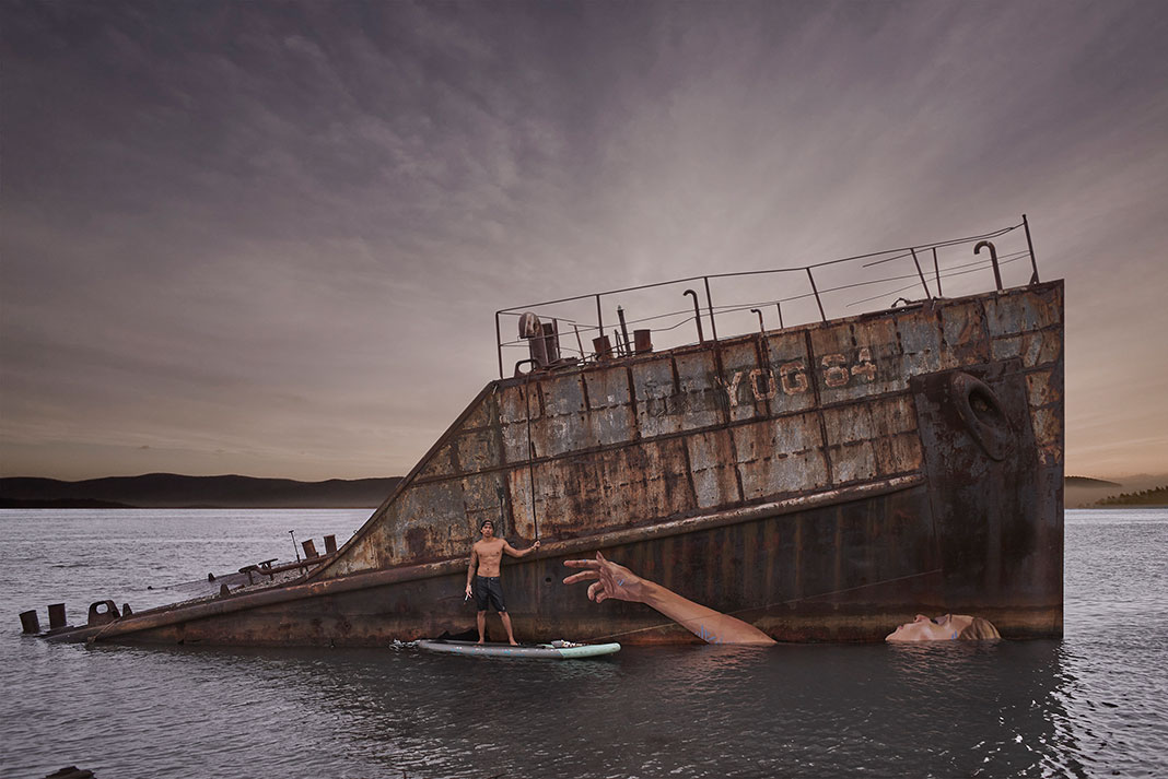 artist Sean Yoro stands on his paddleboard beside a mural he painted on the side of a sunken ship