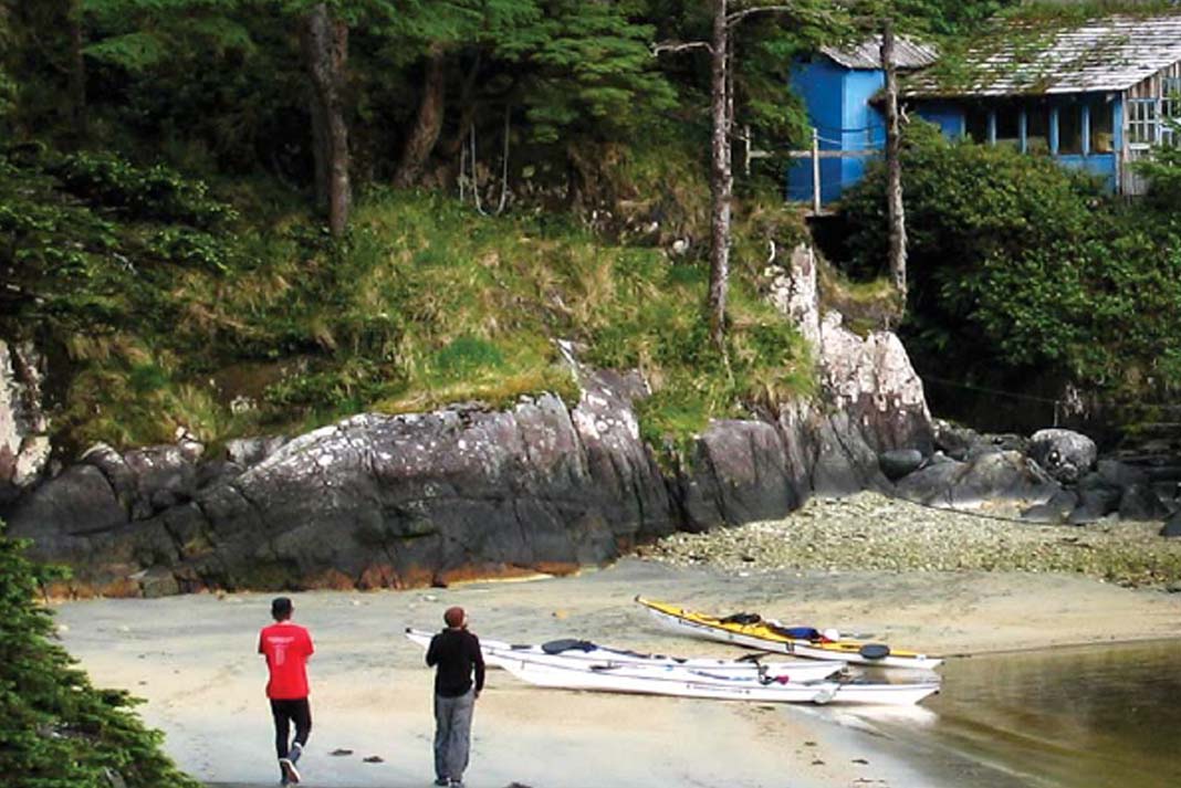 two people walk down a rainforest beach in Haida Gwaii with touring kayaks beached on the sand in front of a small blue building