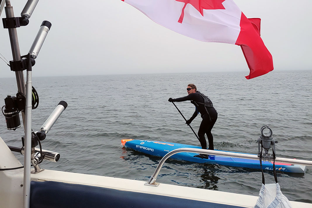 Mike Shoreman paddles beside a boat with Canadian flag during his paddleboarding feat