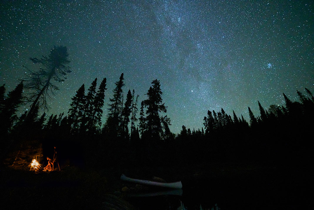 canoe grounded by campsite at night with campfire and stunning milky way stars