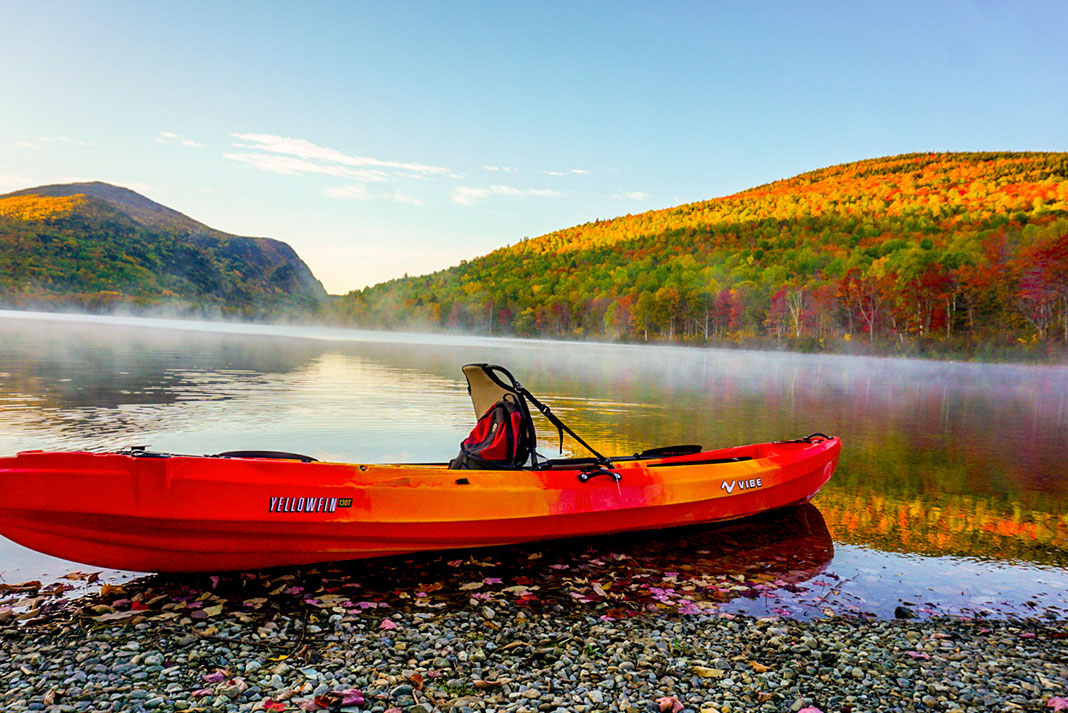 Orange sit-on-top kayak sitting on stony shore with mist over the water and fall colors on the trees in the background