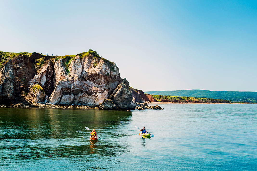 Two kayakers paddle in front of a rocky cliff.
