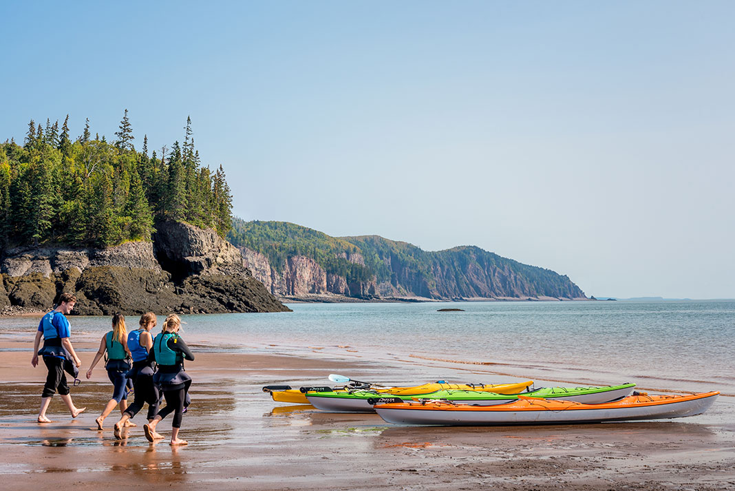 Group of people walk on beach towards sea kayaks with dramatic cliffs along shoreline.