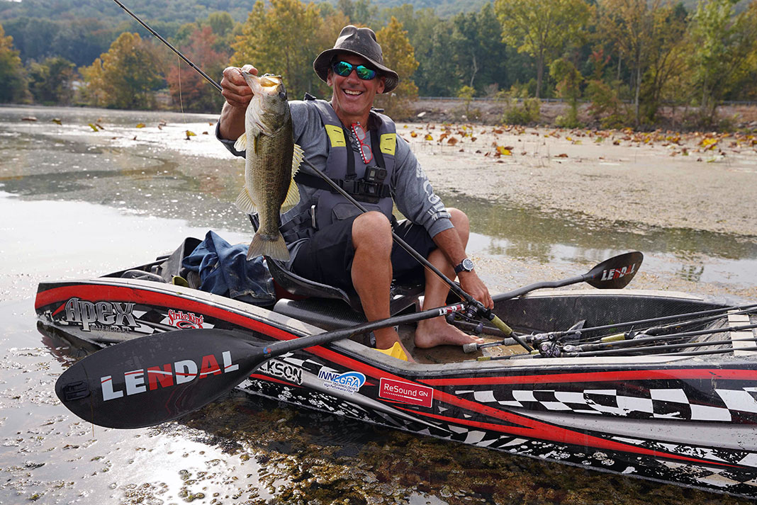 Angler sitting in kayak holds paddle with one hand and a bass with his other. 