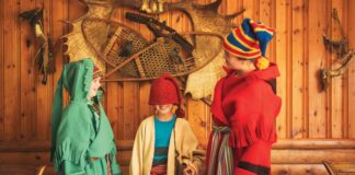 three children inside a wood cabin laugh as they dress up in colorful voyageur sashes