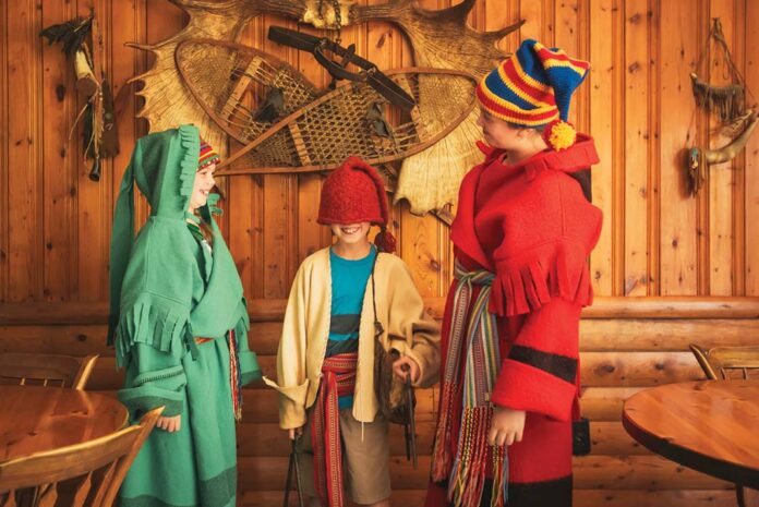 three children inside a wood cabin laugh as they dress up in colorful voyageur sashes