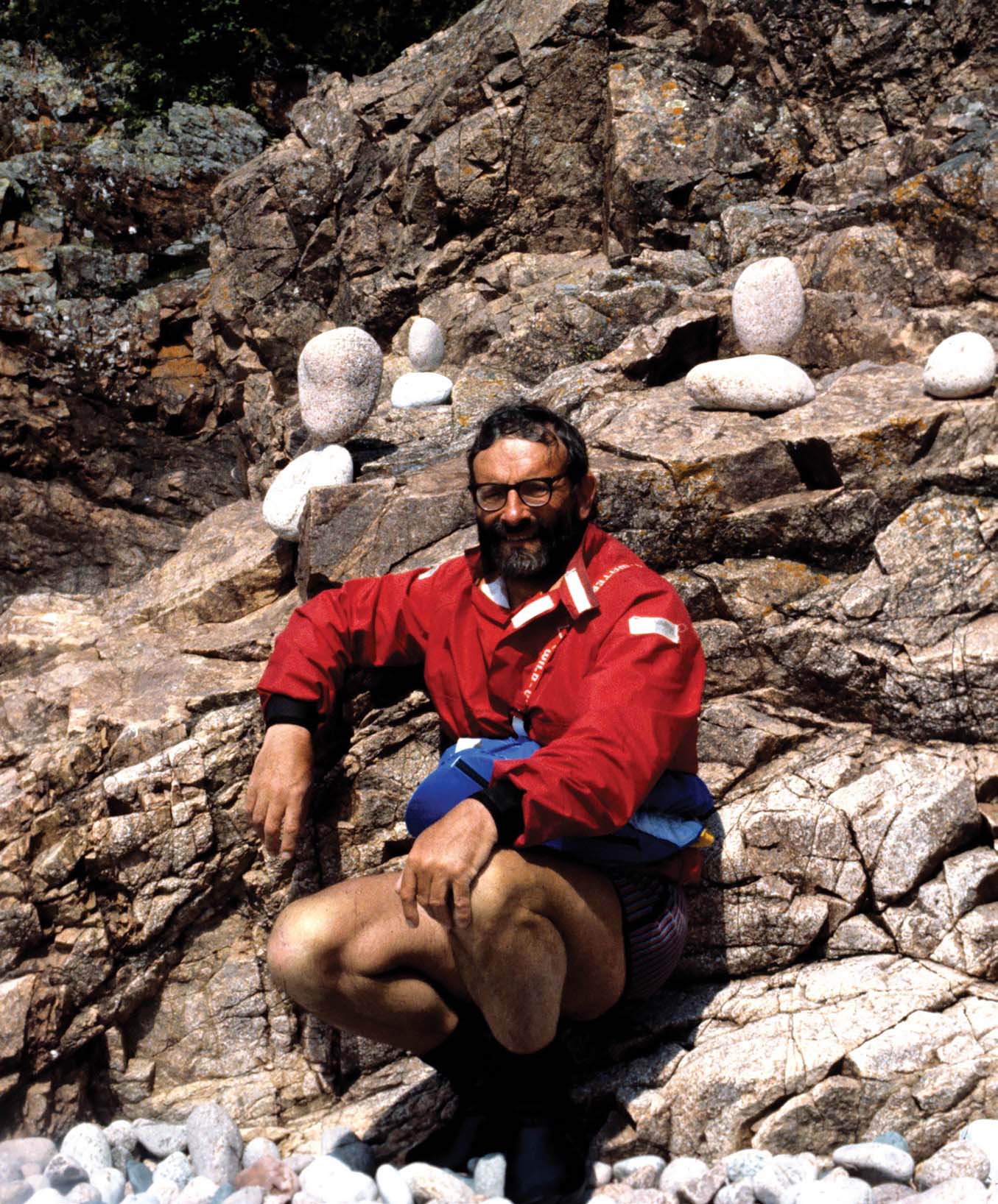 Frank Goodman poses in red drytop on a rocky, pebbly beach