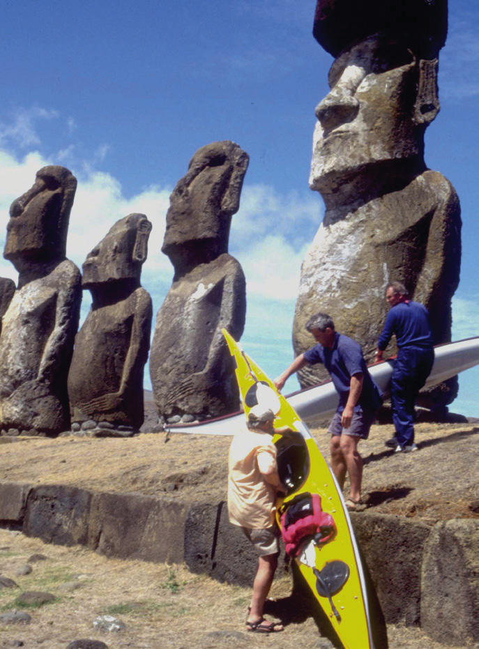 three men carry sea kayaks to pose beside Easter Island statues