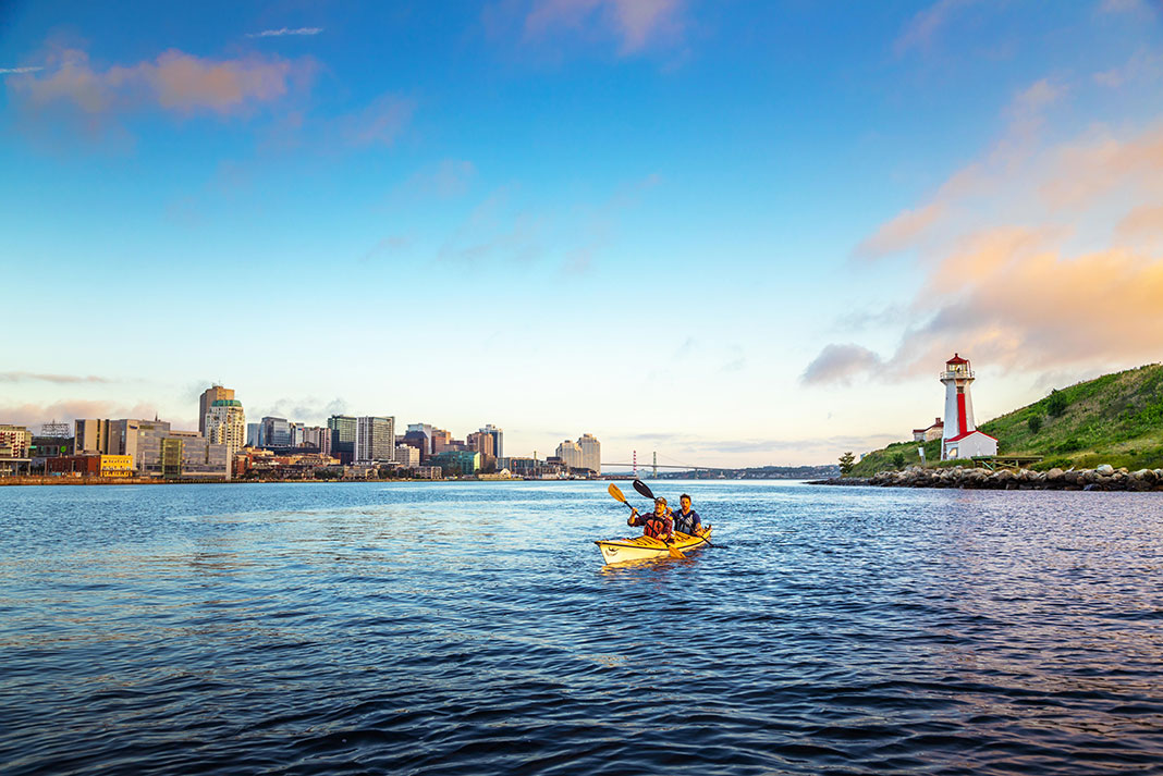Tandem sea kayak being paddled with city buildings on one side and lighthouse on another.