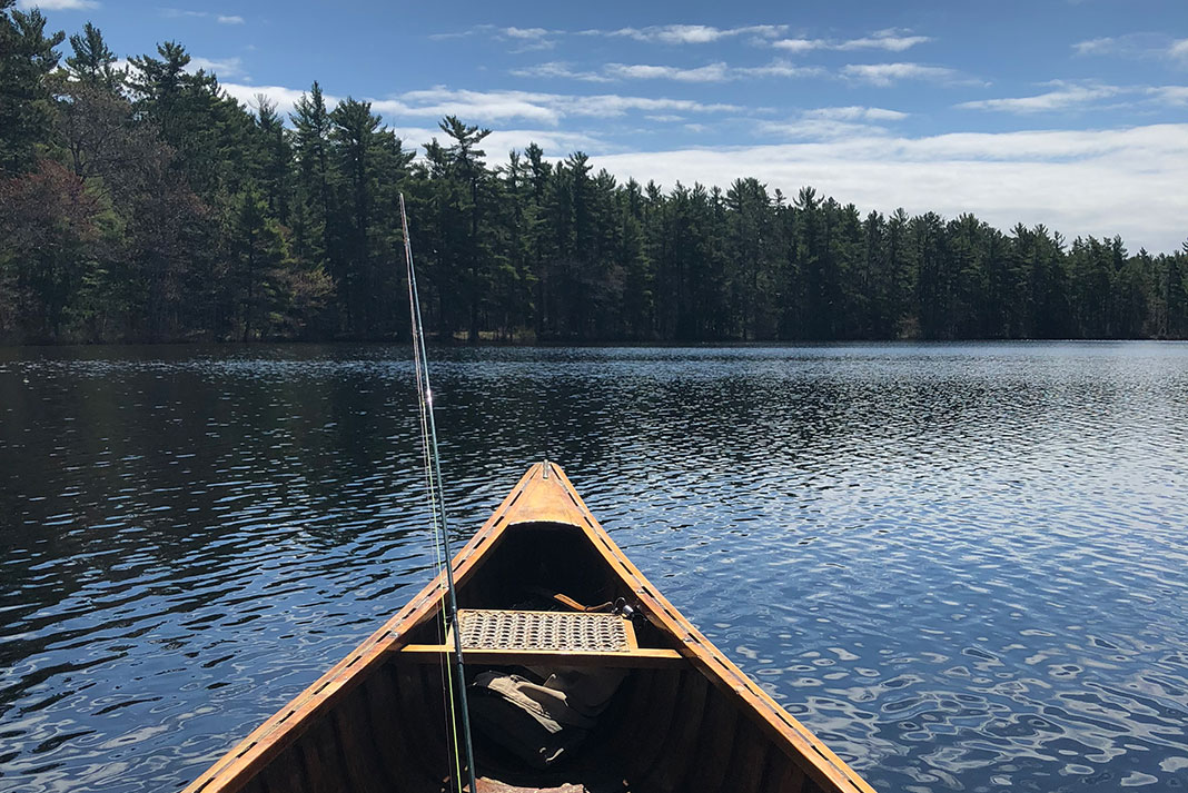 Bow of a wooden canoe and fishing rod looking out over tree-lined lake.