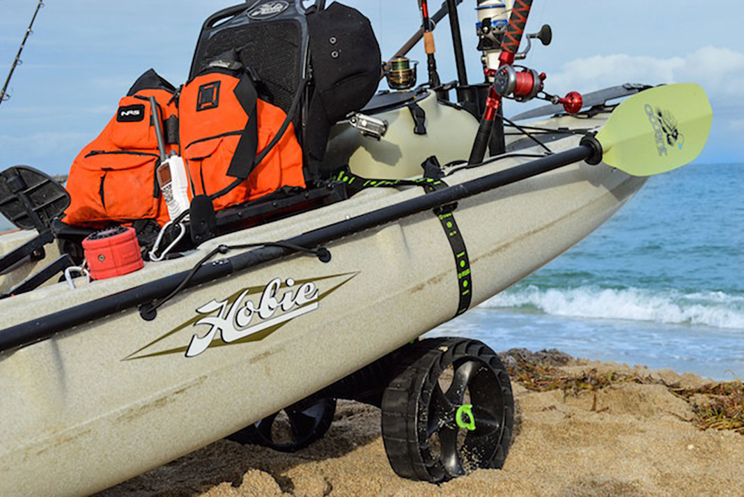 A loaded Hobie kayak sits by the water on a kayak cart with large wheels.