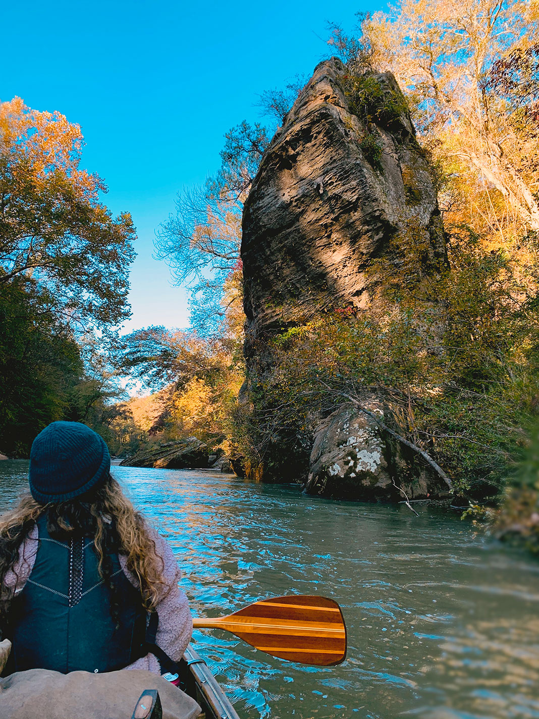Woman sits in bow of canoe holding a paddle and looking up at a rock formation next to the river.