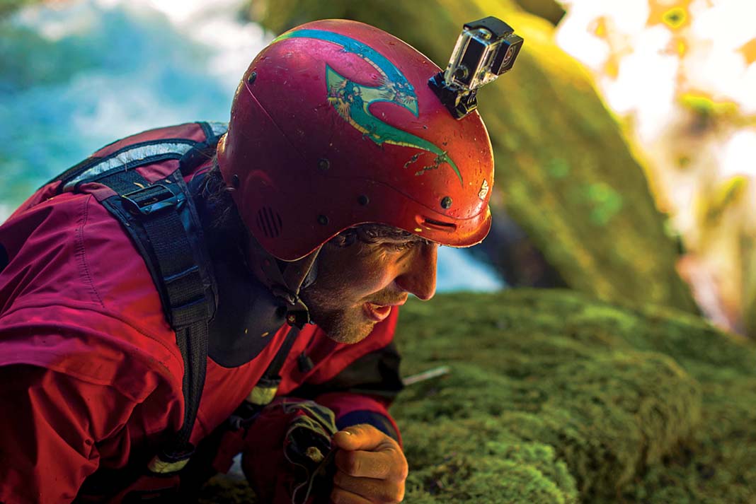 a kayaker in red drysuit and helmet leans on mossy rocks with a helmet-mounted GoPro camera