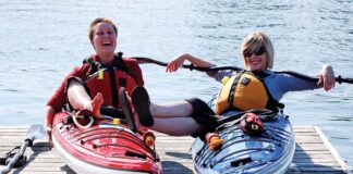 two women lounge on sea kayaks on a dock while laughing