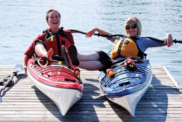 two women lounge on sea kayaks on a dock while laughing