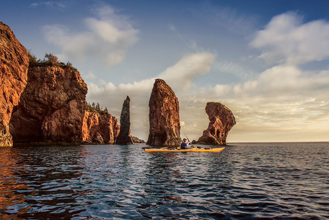 A sea kayaker paddling in front of three towering rock pillars