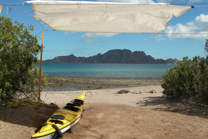 a yellow sea kayak sits on the sand under a tarp rigged for shade with the water and rocky hills of Baja Mexico behind it