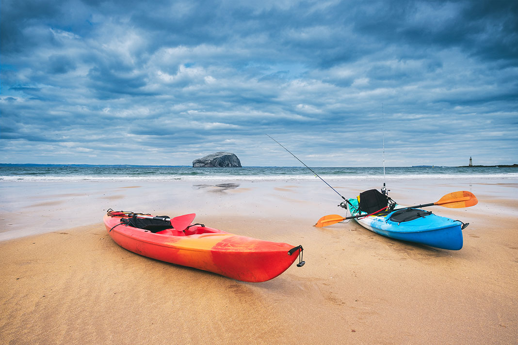 two beginner fishing kayaks sit on a beach in front of cloudy water and lighthouse