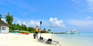 man holds paddle and PFD while standing beside a clear kayak on a tropical white sand beach