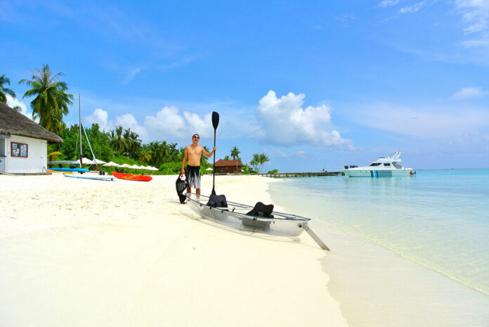 man holds paddle and PFD while standing beside a clear kayak on a tropical white sand beach