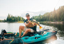 man on a blue Old Town fishing kayak holds up a fish caught while wearing a fishing hat