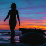 How To Pick The Perfect Women’s Paddleboard woman stands beside paddleboard silhouetted in the sunset