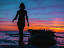 woman stands beside paddleboard silhouetted in the sunset
