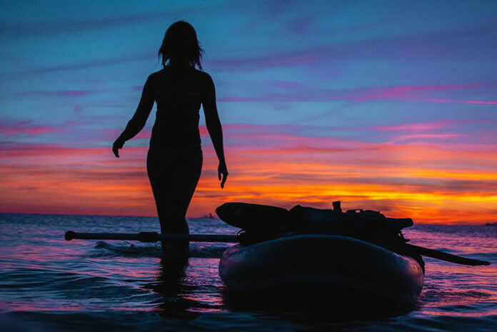 woman stands beside paddleboard silhouetted in the sunset